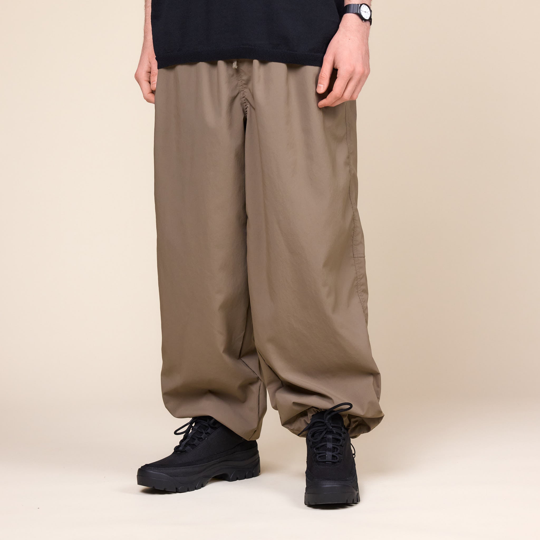 a man wearing a pair of Still by Hand Airy Wide Trousers in a Khaki colour with a pair of black Tarvas shoes on a cream background.