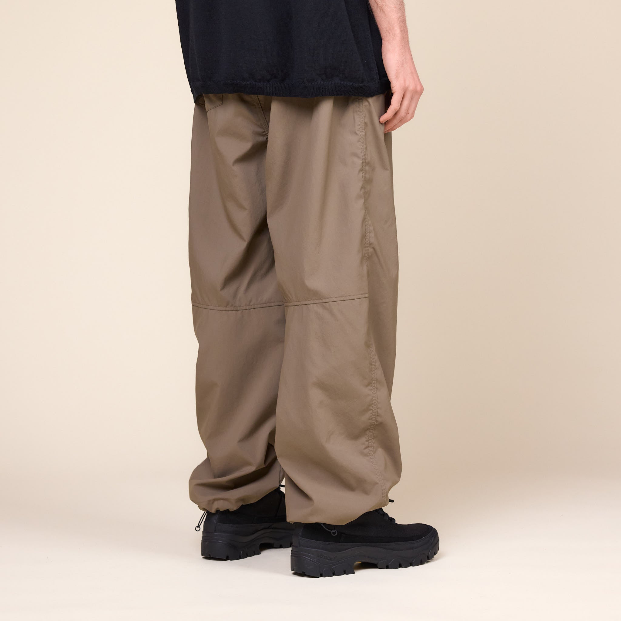 a man wearing a pair of Still by Hand Airy Wide Trousers in a Khaki colour with a pair of black Tarvas shoes on a cream background.