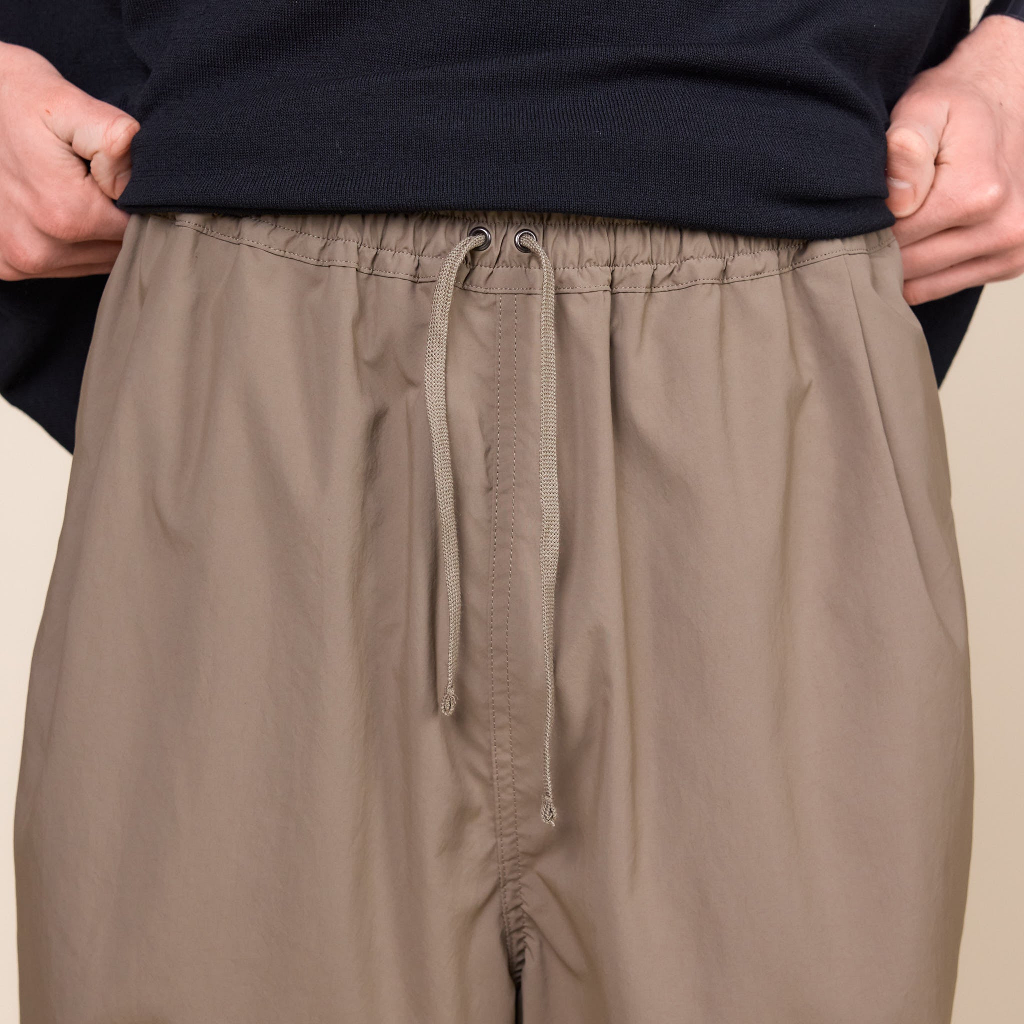 a man showing the waistband of a pair of Still by Hand Airy Wide Trousers in a Khaki colour on a cream background.