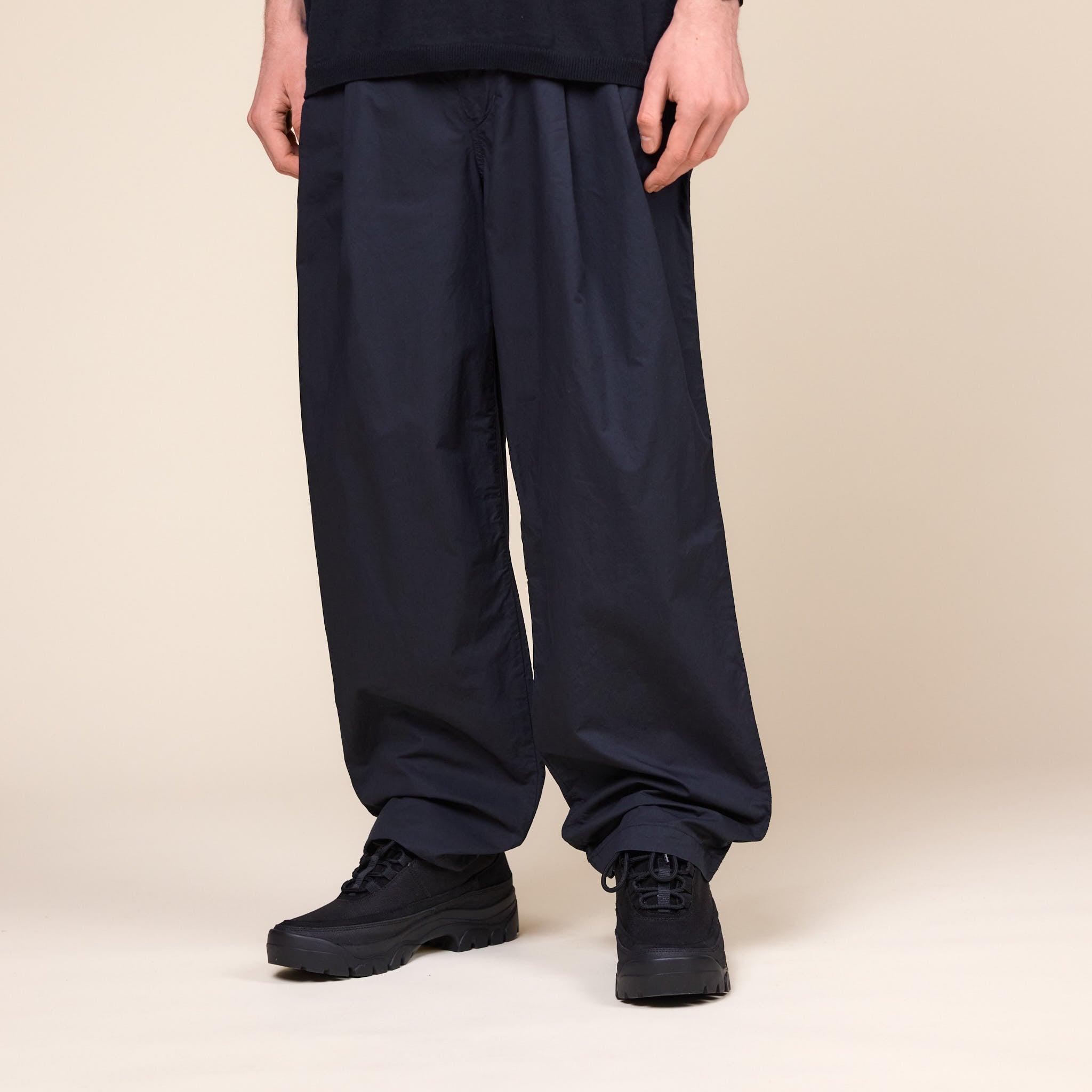 A Man wearing a pair of Still by Hand Garment Dye 2 Tuck Pants in Ink Black colour and a pair of Tarvas Easy Hikes Shoes on a cream background.
