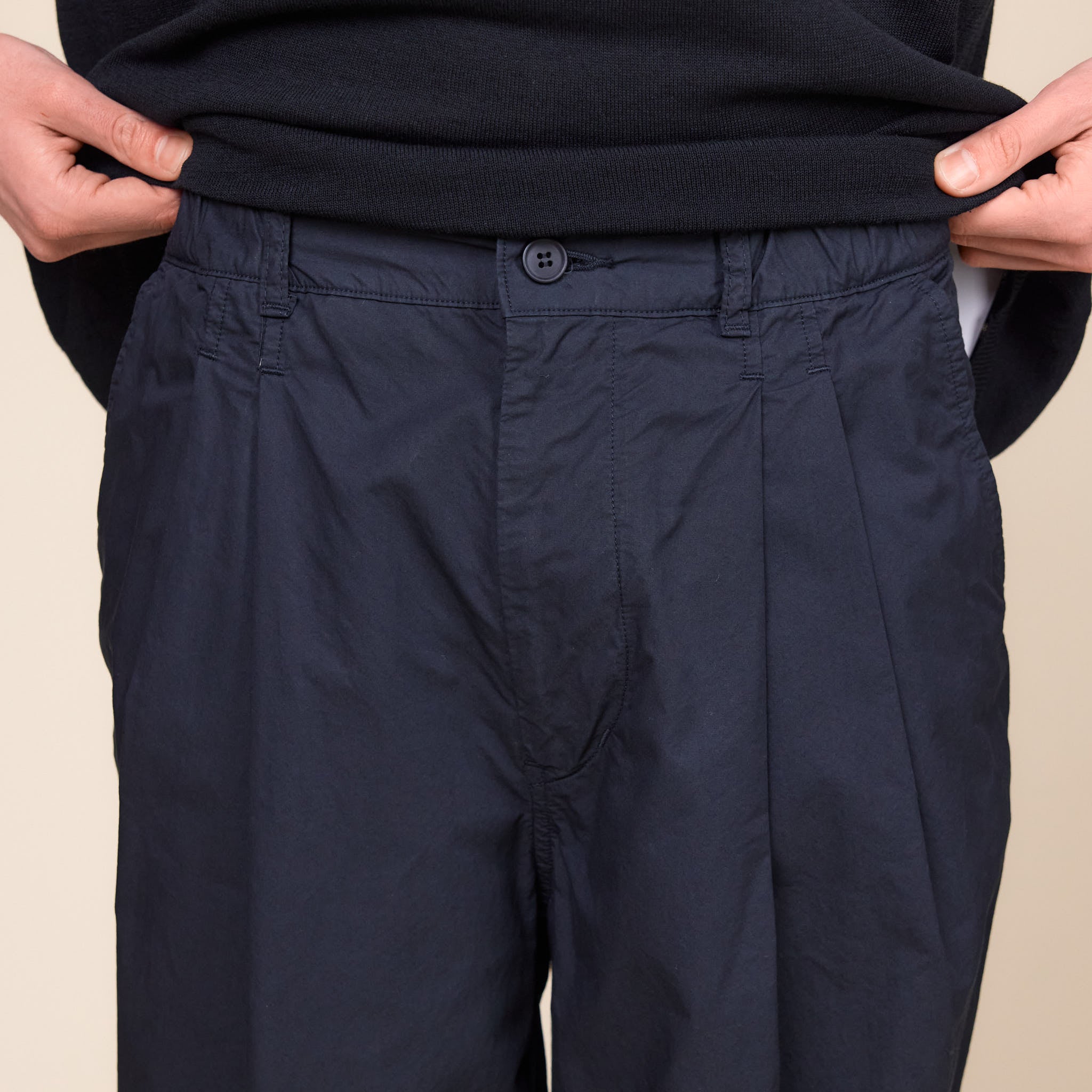 a man showing the waistband on a pair of Still by Hand Garment Dye 2 Tuck Pants in Ink Black colour on a cream background.